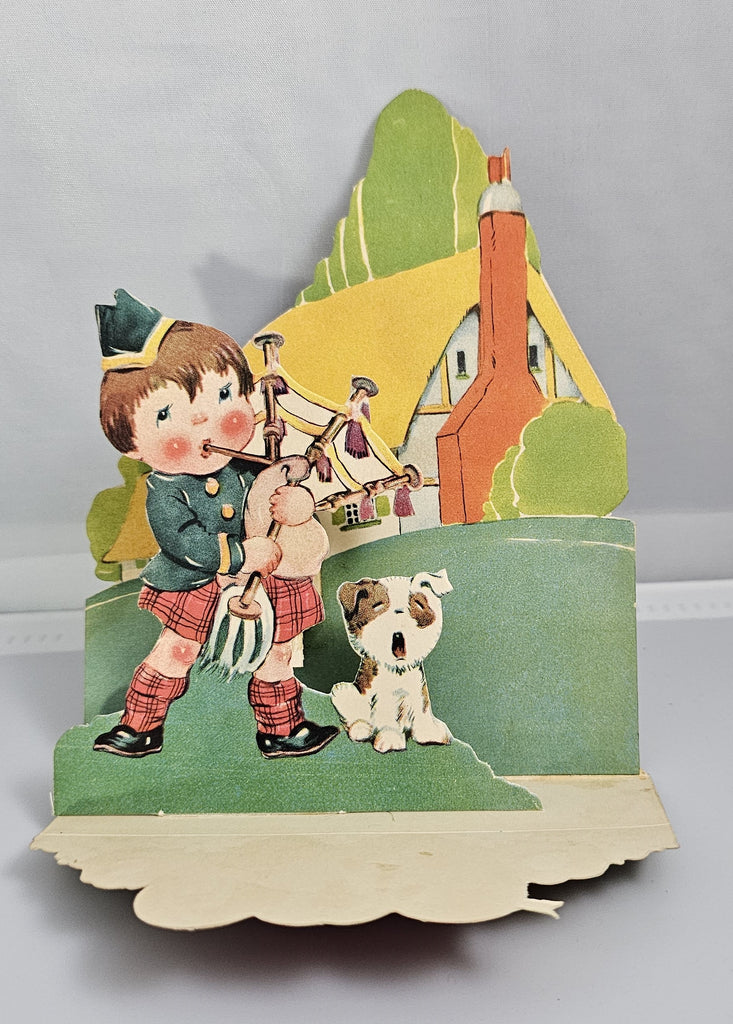 Vintage Die Cut Valentine Little Scottish Boy Playing Bagpipes Outside a Cottage with Hearts and Puppy Dog