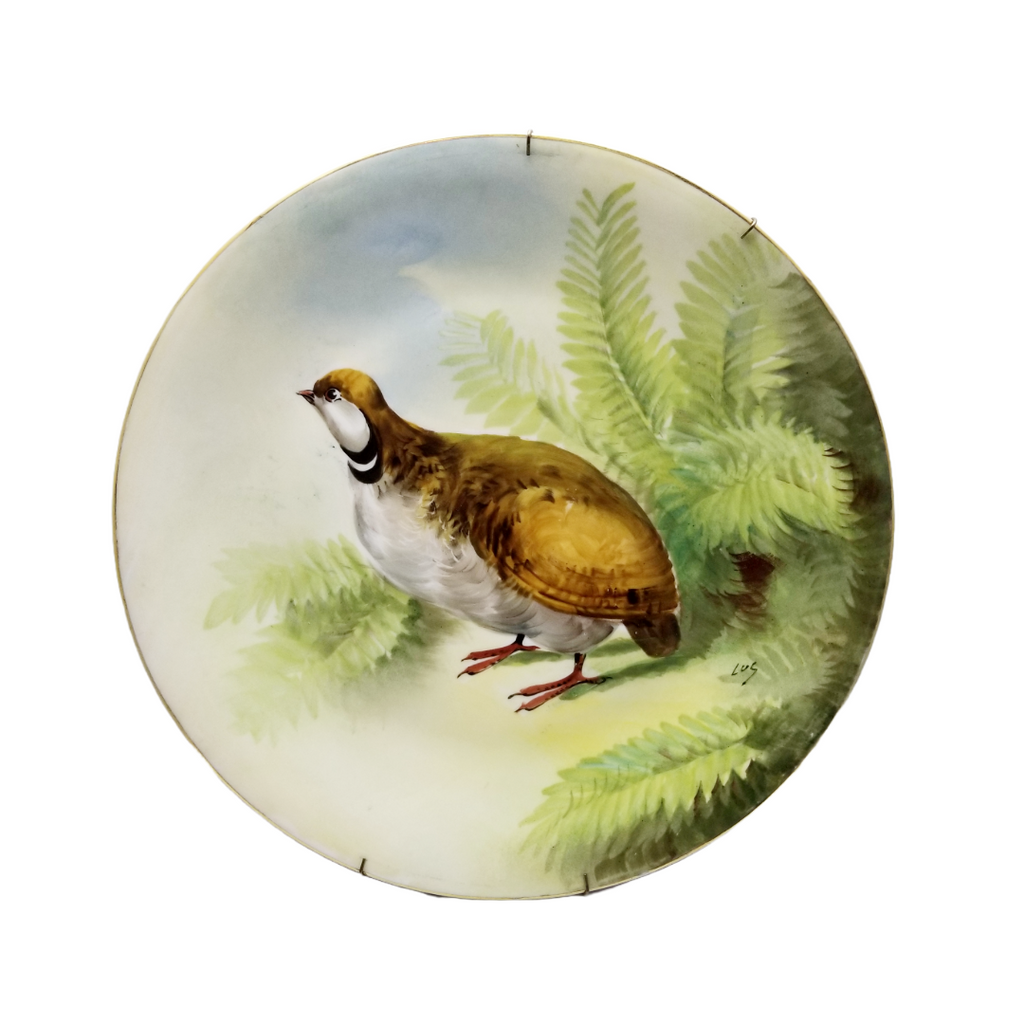 A Limoges porcelain round charger plate with a hand-painted artist-signed quail in the center, surrounded by green foliage and a blue sky background.